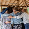 Groep volwassenen in groepsomhelzing tijdens ondersteunende sessie in rieten paviljoen, sfeer passend bij ambulante of residentiële eetstoornis behandeling van BeLeef.
