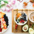 Houten snijplank met kom yoghurt met bessen en kersen, kop koffie, halve avocado; links bord met kleurrijke fruit- en groenteschijfjes op witte tafel naast tulpen.