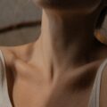 Nek en sleutelbeenderen van persoon in witte spaghettitop, intieme close-up, zacht schuin belicht, weinig achtergronddetails.